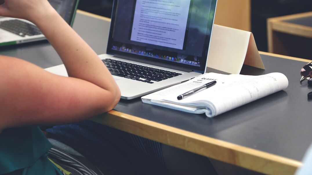 Picture of a woman's arm. She is leaning on a table. There is an open laptop on the table and a notebook beside the laptop.